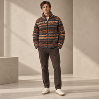 A man wearing a patterned zip-up fleece jacket with horizontal multicolor stripes, dark brown pants, and white sneakers, standing in a minimalist beige studio setting.