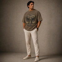 Man wearing a gray t-shirt with 'Freedom Vintage' text and white pants against a brown background