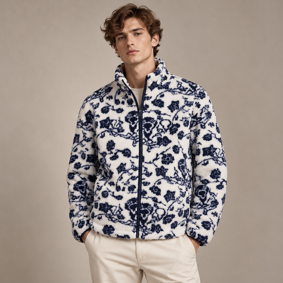A man wearing a white sherpa fleece jacket with a navy blue floral pattern, paired with cream-colored trousers, standing against a solid beige studio background.