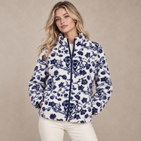 A woman modeling a white sherpa fleece jacket with a navy blue floral toile pattern, paired with cream-colored pants, against a neutral beige studio background.