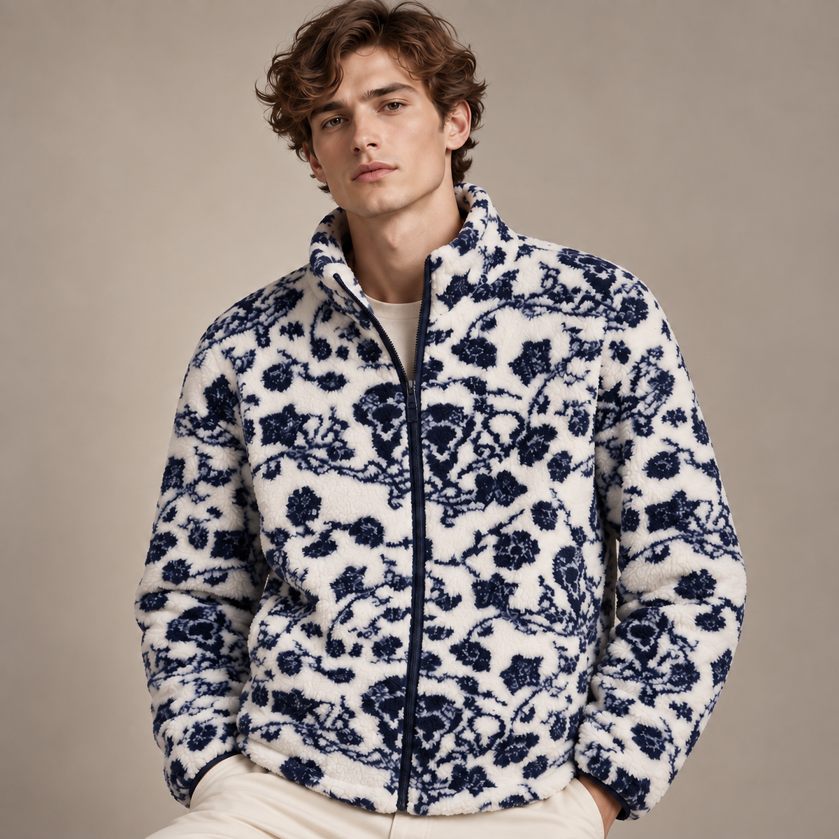 A man wearing a white sherpa fleece zip-up jacket with a navy blue floral pattern, paired with cream pants, set against a solid beige studio background.