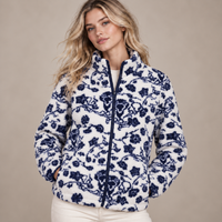 A woman wearing a white sherpa-style zip-up jacket with a navy blue floral pattern, paired with cream pants, set against a plain beige studio background.