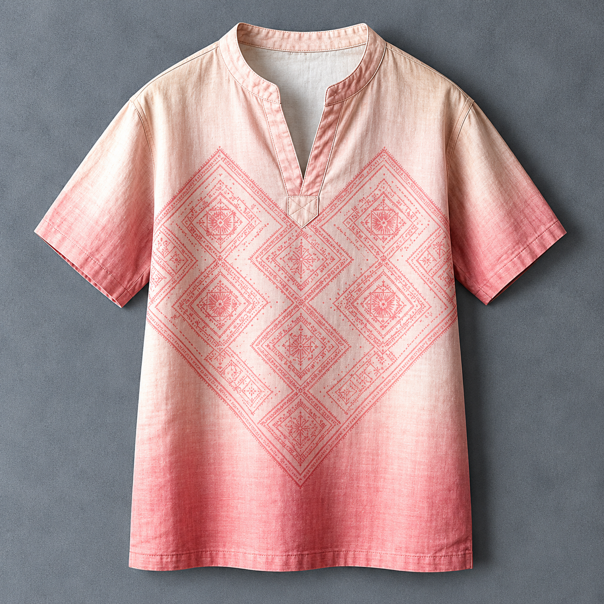 A short-sleeve V-neck tunic top with a pink and beige gradient and a large geometric diamond pattern printed across the front, laid flat against a dark gray background.