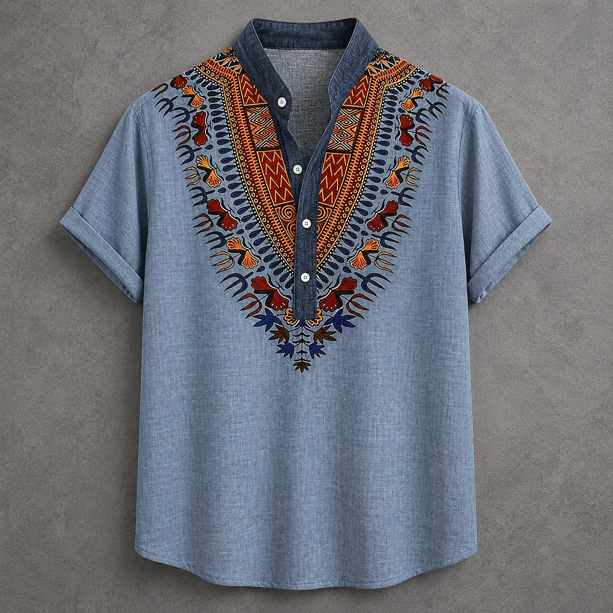 A light blue short-sleeve button-up shirt with a dark blue collar and a vibrant, multicolored African dashiki-style embroidered yoke on the chest, displayed flat against a textured gray background.