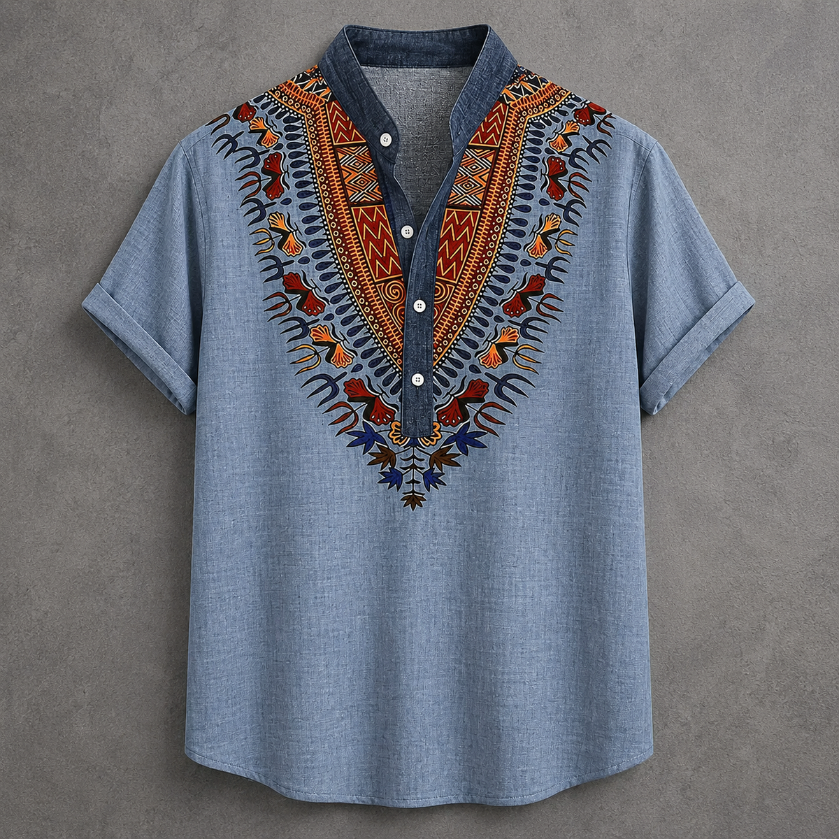 A light blue short-sleeve button-up shirt with a dark blue collar and a vibrant, multicolored African dashiki-style embroidered yoke on the chest, displayed flat against a textured gray background.