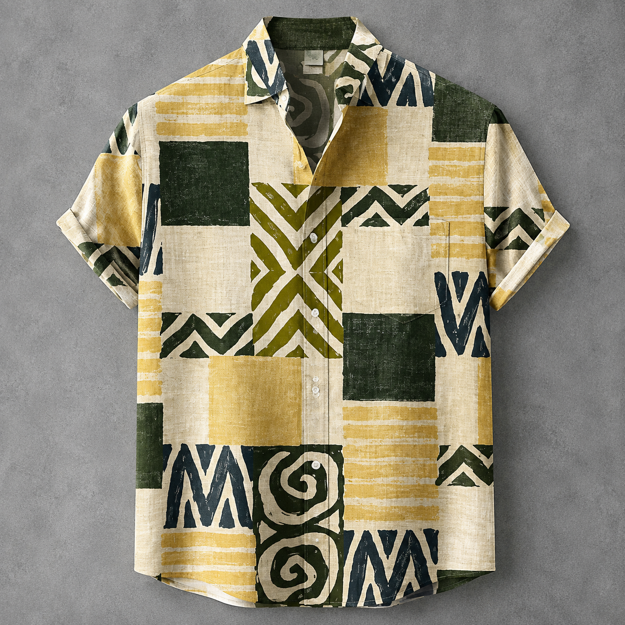 A short-sleeve button-up shirt with a patchwork geometric print in olive green, mustard yellow, cream, and dark navy, laid flat on a textured gray background.