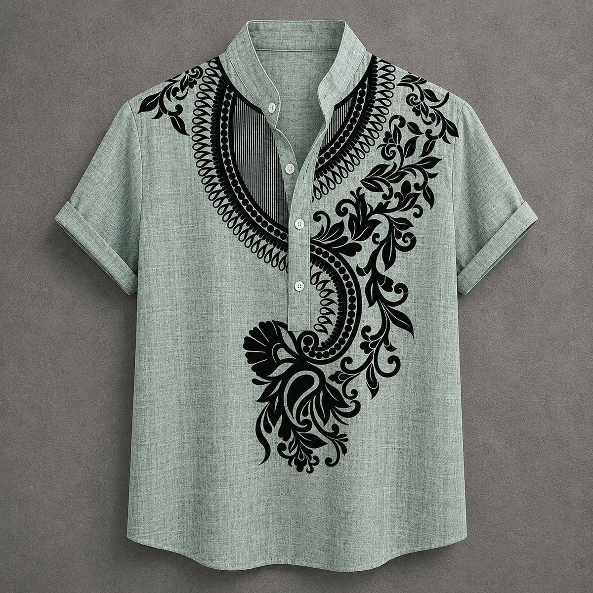A short-sleeve button-up shirt in a light sage green linen-like fabric, featuring a large black ornate floral and scroll pattern printed or embroidered across the chest and collar area. The shirt is displayed flat against a textured gray background.