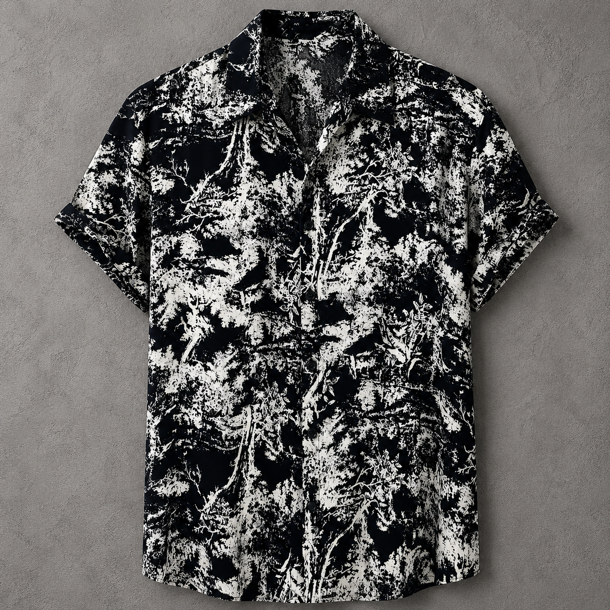 A short-sleeve button-up camp shirt with a black and white abstract leafy pattern, laid flat on a textured gray background.