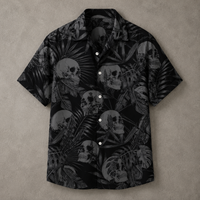 A short-sleeve button-up camp shirt with a black and gray tropical print featuring skulls and palm leaves, laid flat on a textured gray background.