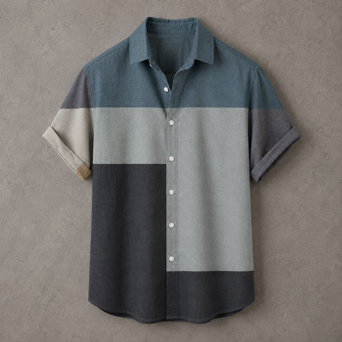 A short-sleeve button-up shirt with a color-blocked design in slate blue, light gray, dark charcoal, and beige, laid flat against a textured gray background.