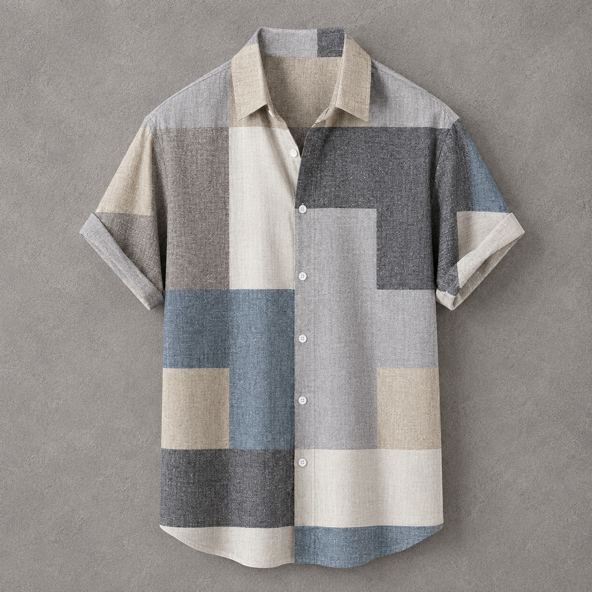 A short-sleeve button-up shirt with a geometric patchwork pattern in beige, gray, blue, and white, displayed flat against a textured gray background.