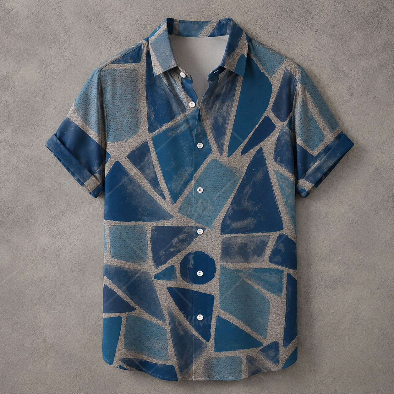 A short-sleeve button-up shirt with a blue and gray geometric mosaic pattern, laid flat on a textured gray background.