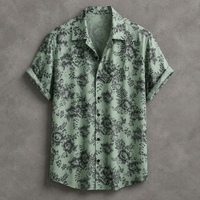 A short-sleeve button-up camp shirt in sage green with a dark gray floral pattern, laid flat on a textured gray background.