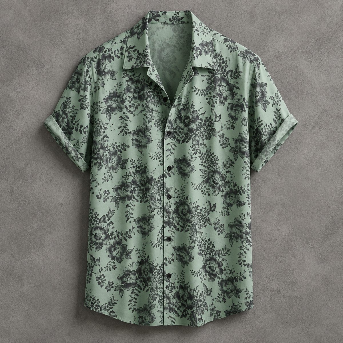 A short-sleeve button-up camp shirt in sage green with a dark gray floral pattern, laid flat on a textured gray background.