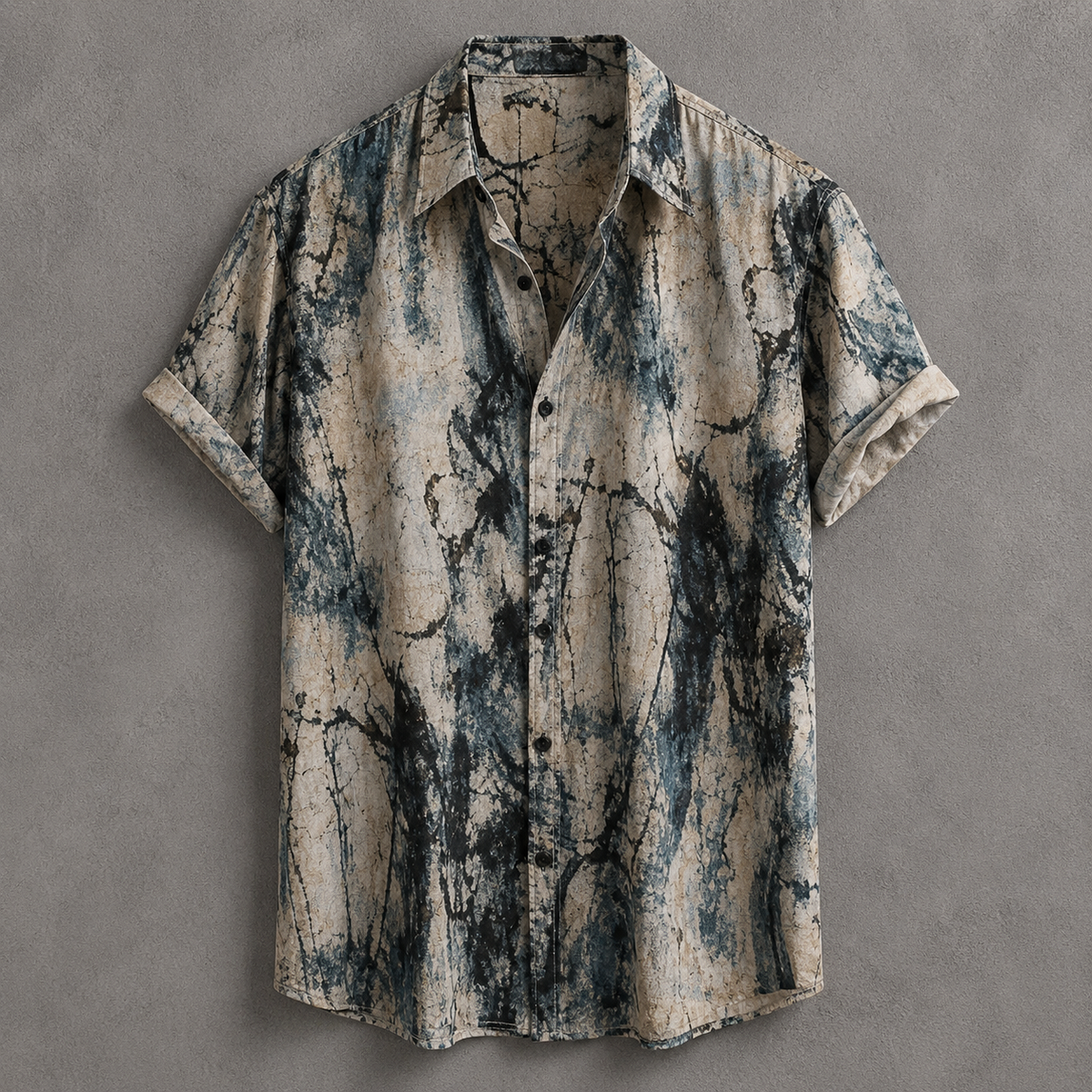 A short-sleeve button-up shirt with a distressed, abstract tie-dye or marble-like print in beige, slate blue, and charcoal gray, laid flat against a textured gray background.