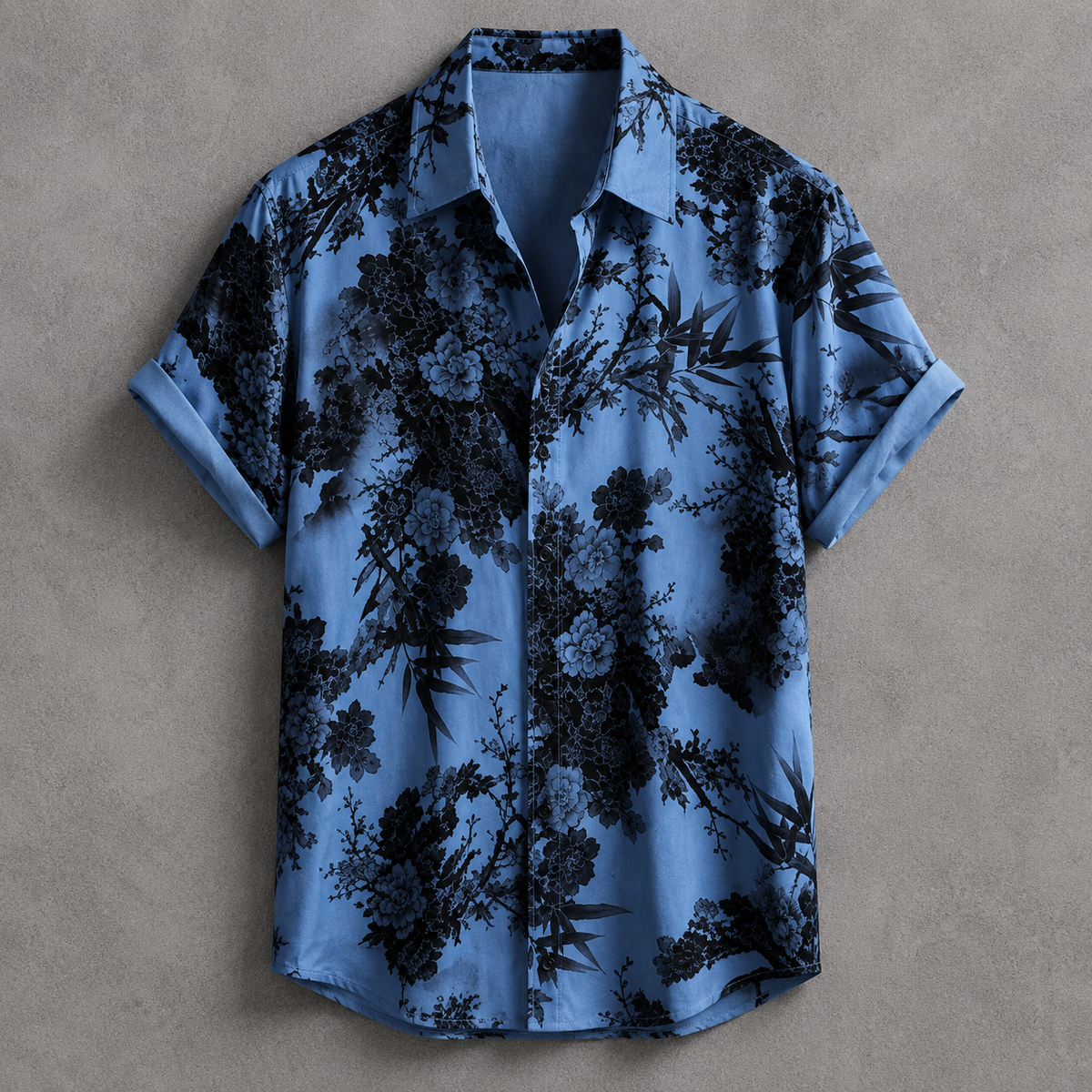 A short-sleeve button-up shirt in light blue with a black floral and botanical print, laid flat against a textured gray background.