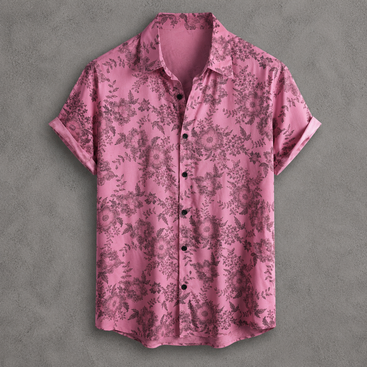 A short-sleeve button-up shirt in pink with a dark floral and botanical pattern, laid flat against a textured gray background.