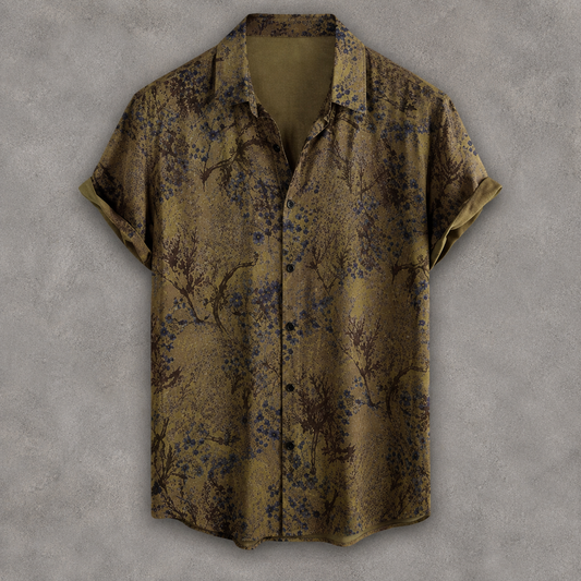 A short-sleeve button-up shirt with a muted olive-brown base and an allover abstract floral or branch-like print in dark blue and black, displayed against a textured gray background.