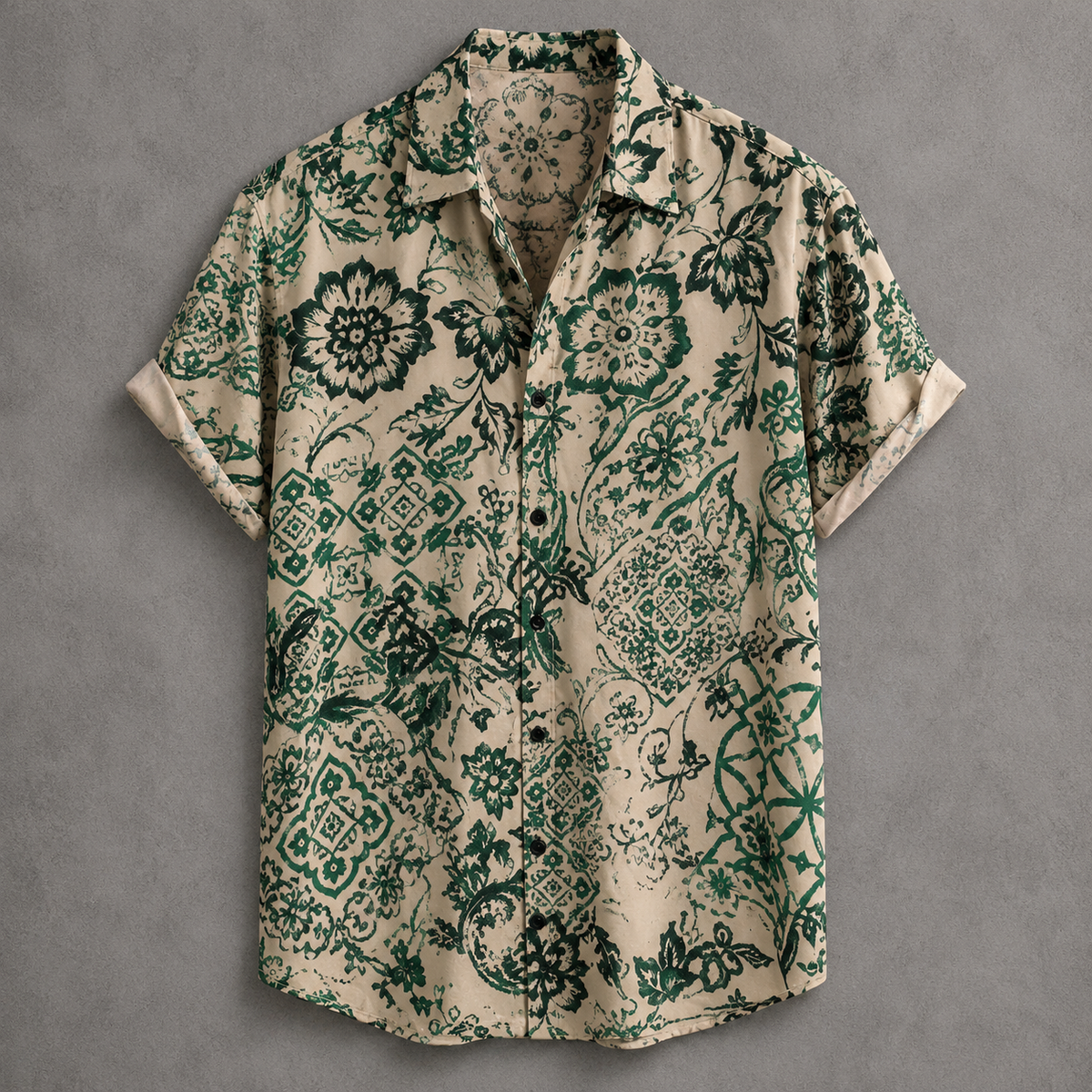 A short-sleeve button-up shirt with a beige base and an intricate green floral and paisley pattern, laid flat against a textured gray background.