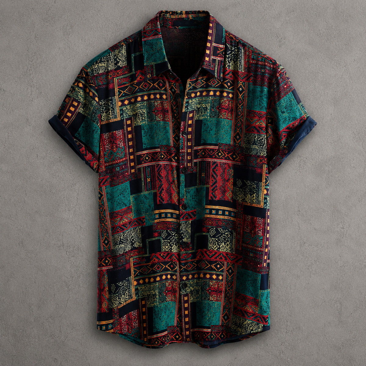 A short-sleeve button-up shirt with a complex geometric patchwork pattern in teal, red, black, and gold, laid flat against a textured gray background.