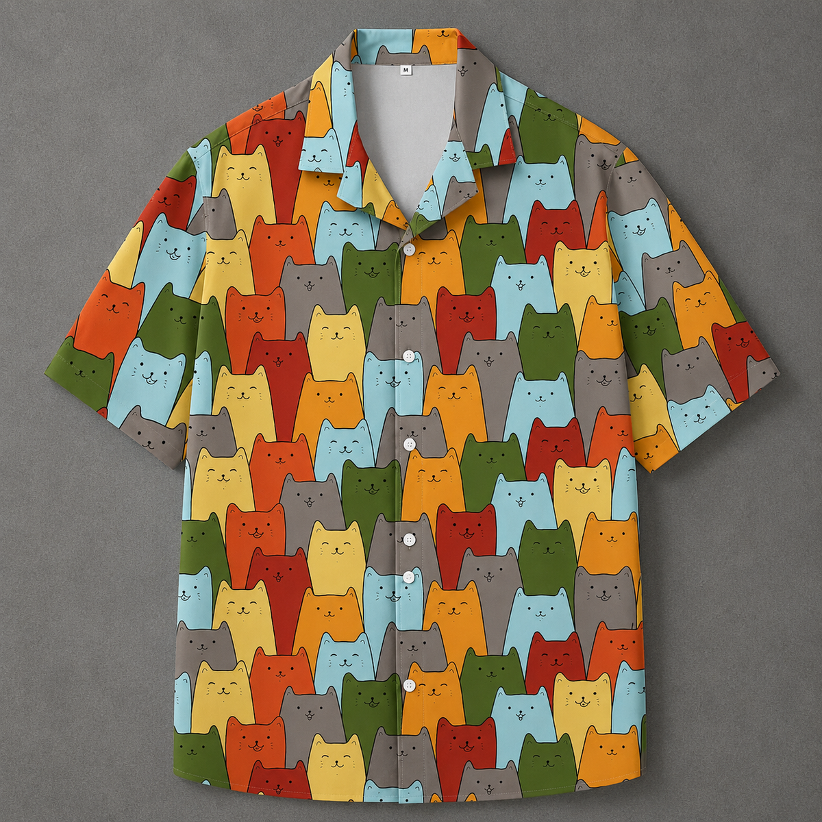 A short-sleeve button-up shirt with an all-over repeating pattern of cartoon cat faces in multiple colors, including orange, yellow, red, green, blue, and gray, laid flat on a dark gray background.