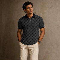 Man wearing a black patterned polo shirt and beige pants against a brown background