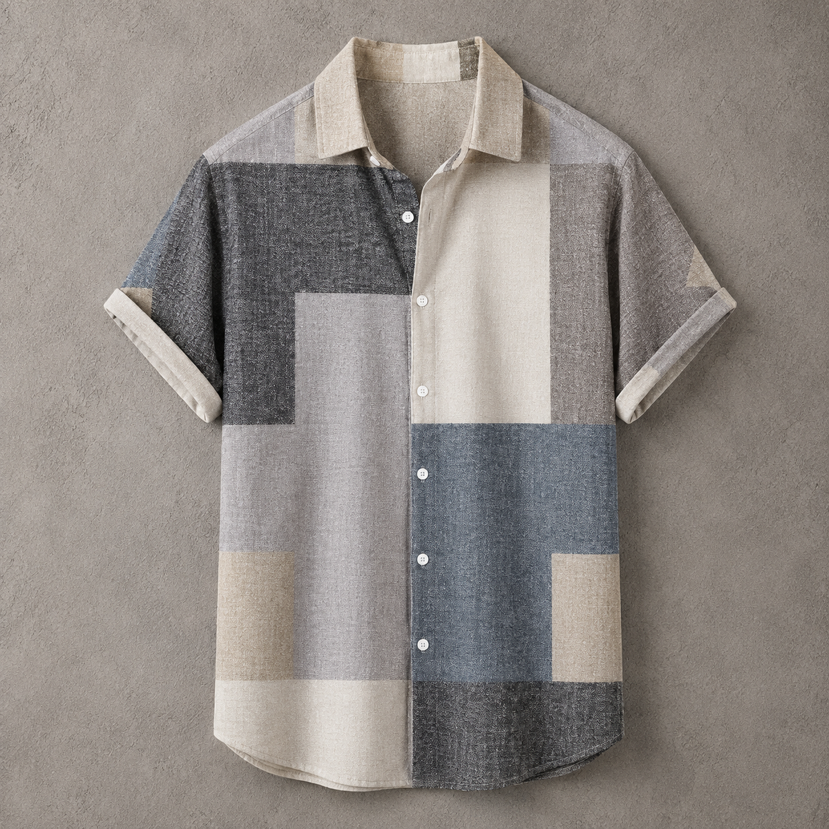 A short-sleeve button-up shirt with a geometric patchwork pattern in beige, gray, and slate blue, laid flat against a textured gray background.