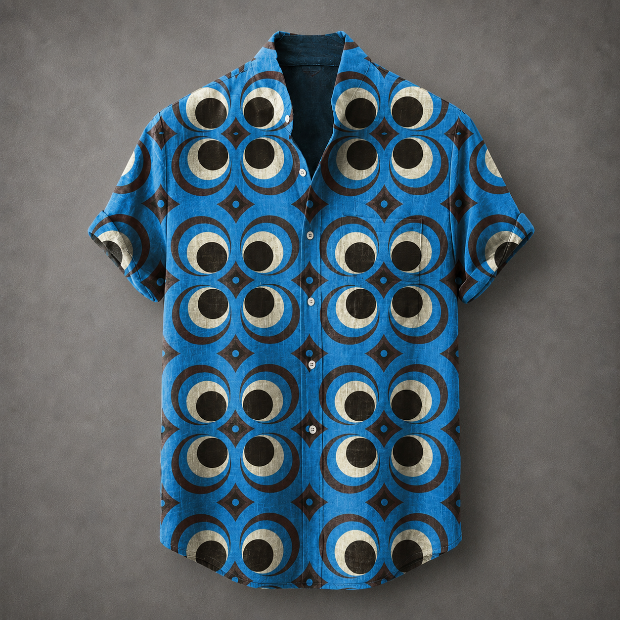 A short-sleeve button-up shirt with a bold blue, black, and beige geometric eye pattern, laid flat against a textured gray background.