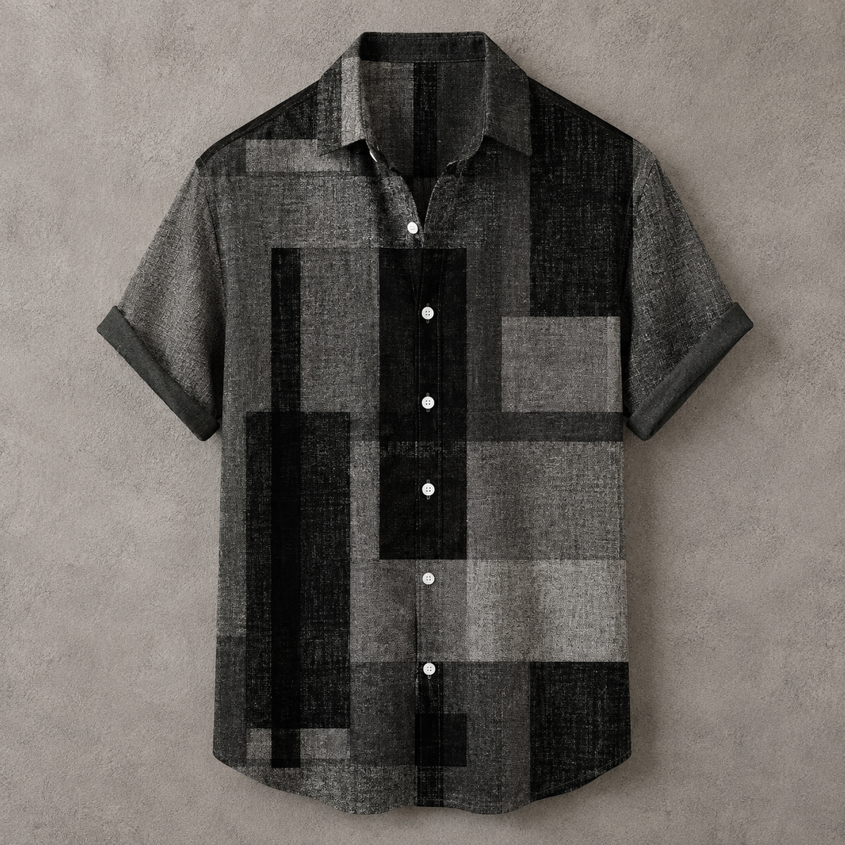 A short-sleeve button-up shirt with a geometric block pattern in black, gray, and charcoal, laid flat against a textured gray background.