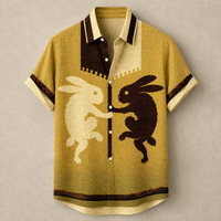 A short-sleeve button-up shirt in textured mustard yellow fabric, featuring a split collar in cream and dark brown, with a central graphic of a cream rabbit facing a dark brown rabbit. The shirt has dark brown trim along the sleeve cuffs and hem.