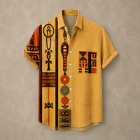 A short-sleeve button-up shirt in mustard yellow with a bold African tribal-inspired geometric and symbolic print in brown, black, orange, and beige, displayed against a textured gray background.