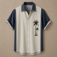 A short-sleeve button-up shirt with a beige body, navy blue collar and sleeve panels, and a black palm tree graphic on the left chest, displayed against a solid tan background.
