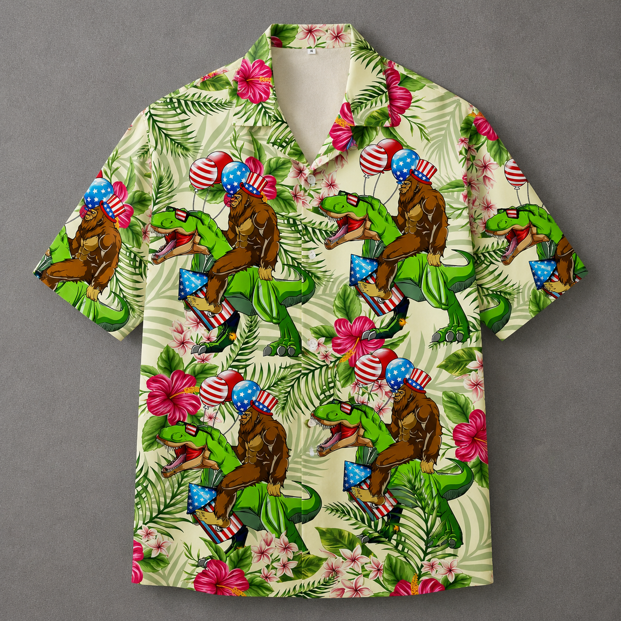 A short-sleeve camp collar button-up shirt with a vibrant, all-over graphic print. The pattern features green dinosaurs riding brown gorillas, decorated with American flags, set against a tropical background of palm leaves, pink hibiscus flowers, and blue-and-white flags, all on a light cream base.