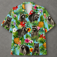 A short-sleeve button-up camp shirt with a vibrant tropical print featuring raccoons, pineapples, palm leaves, and hibiscus flowers on a light blue background, displayed flat against a gray backdrop.