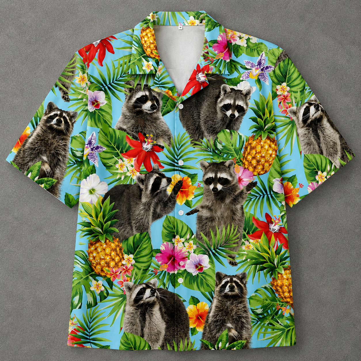 A short-sleeve button-up camp shirt with a vibrant tropical print featuring raccoons, pineapples, palm leaves, and hibiscus flowers on a light blue background, displayed flat against a gray backdrop.