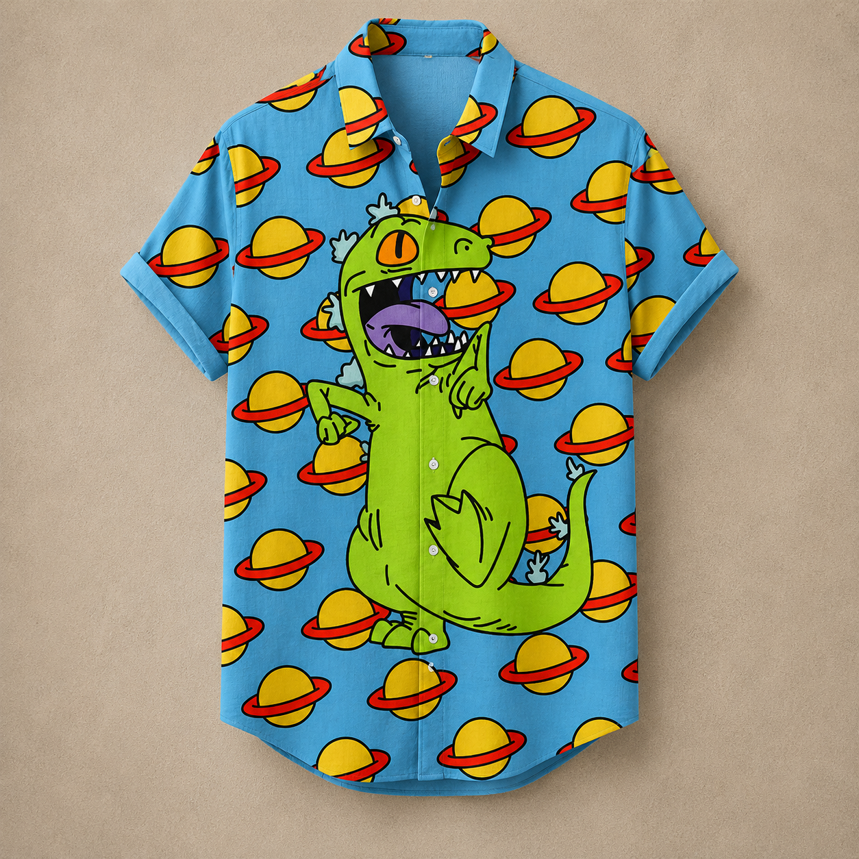 A short-sleeve button-up shirt in bright blue, featuring a cartoon green dinosaur with a gold crown and a repeating pattern of yellow planets with red rings, laid flat on a beige background.