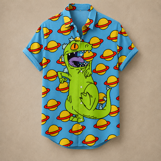 A short-sleeve button-up shirt in bright blue, featuring a cartoon green dinosaur with a gold crown and a repeating pattern of yellow planets with red rings, laid flat on a beige background.