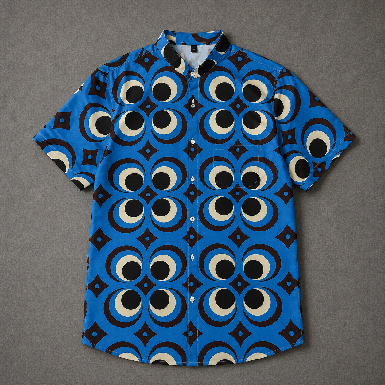 A short-sleeve button-up shirt with a vibrant blue base and a bold geometric pattern of large circular eyes and black diamond shapes, laid flat on a textured gray background.