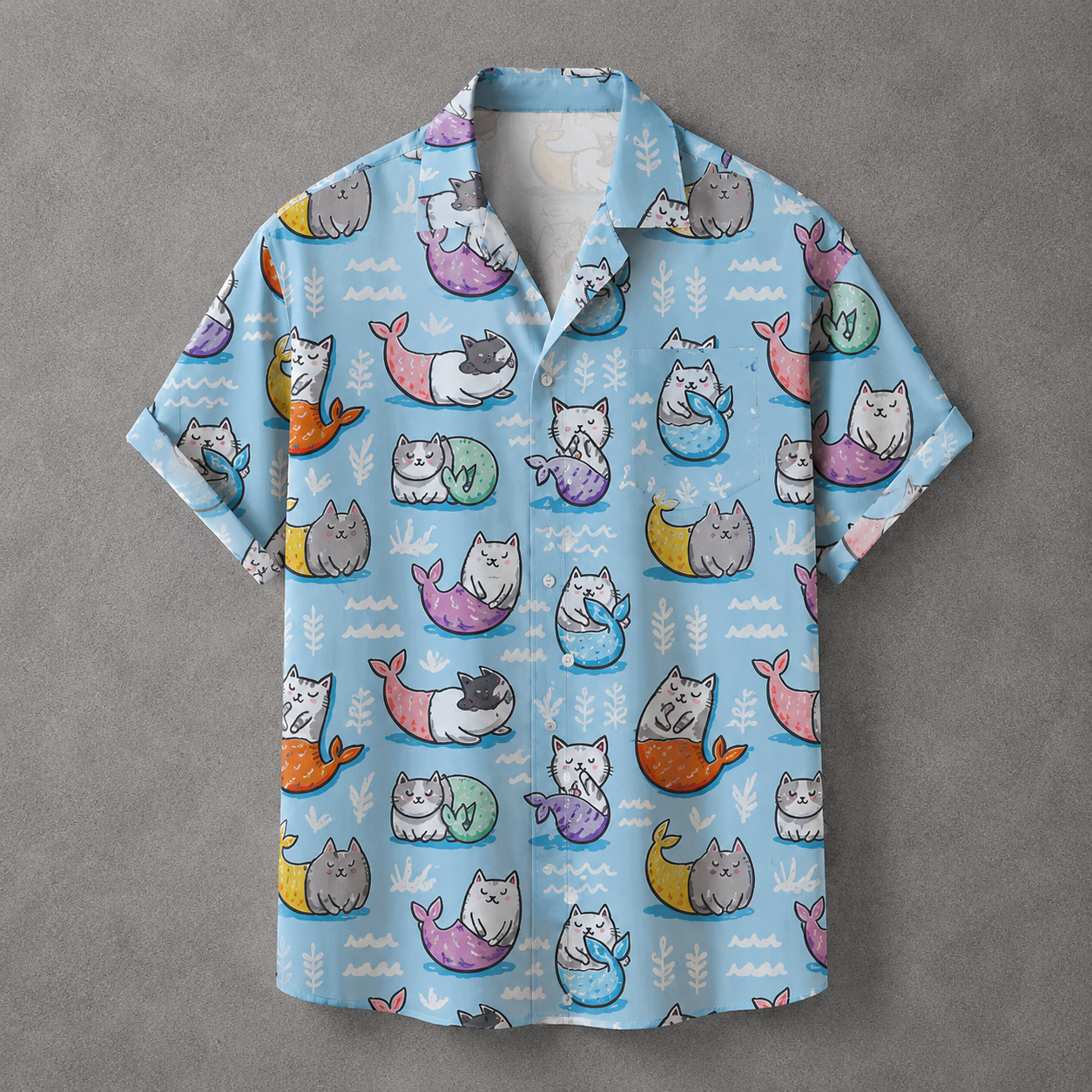 A light blue short-sleeve button-up shirt with an all-over print of cartoon mermaids in pastel colors, laid flat on a textured gray background.