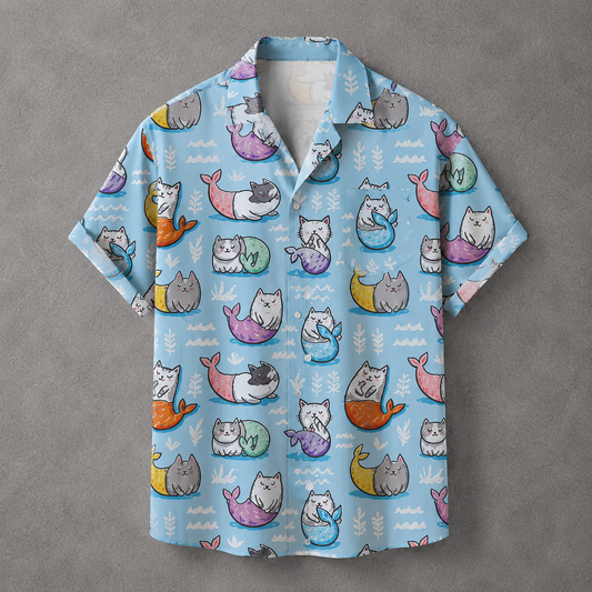 A light blue short-sleeve button-up shirt with an all-over print of cartoon mermaids in pastel colors, laid flat on a textured gray background.