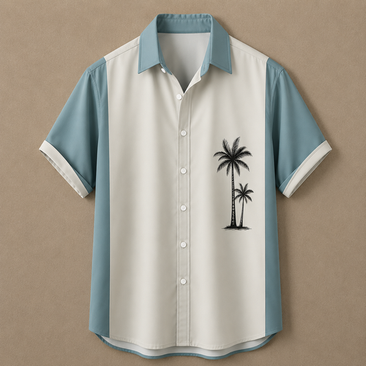 A short-sleeve button-up shirt with a color-blocked design, featuring a cream body, light blue shoulders and collar, and a black palm tree graphic on the left chest, displayed against a textured tan background.
